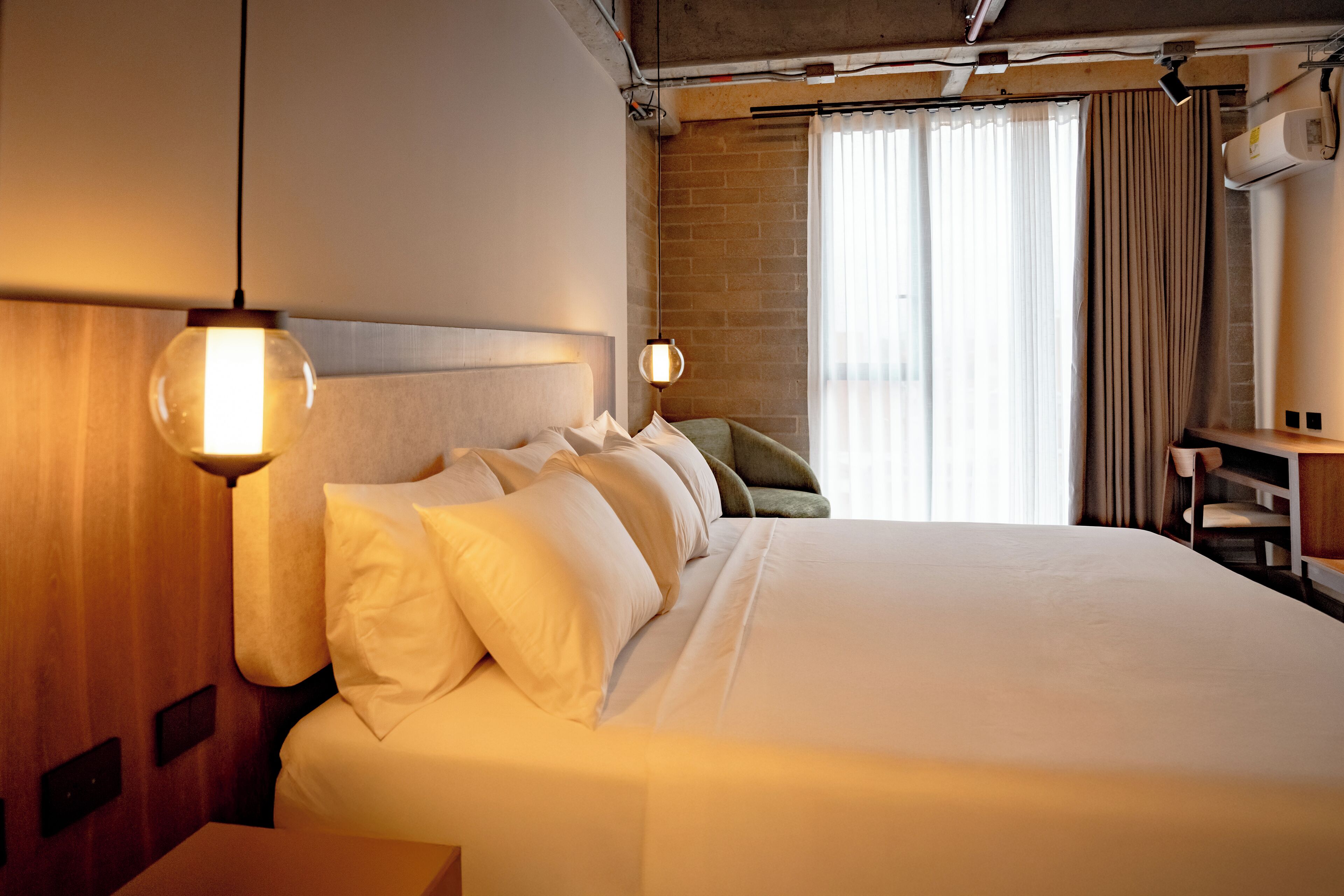 A hotel room with a large bed, two bedside lamps, a wooden headboard, a brick wall, and a window with curtains.