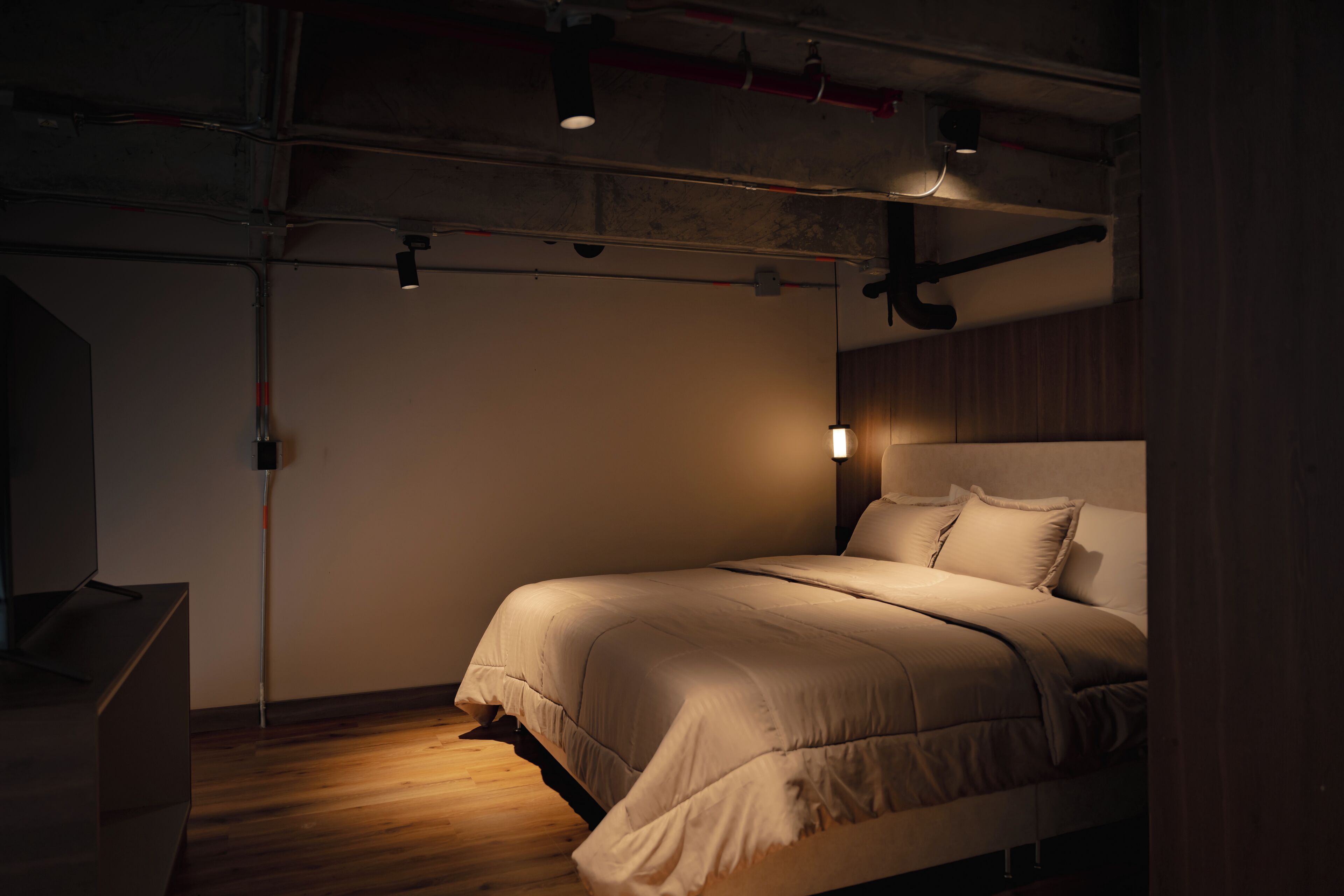 Un dormitorio moderno con cama, suelo de madera y tuberías a la vista en el techo.