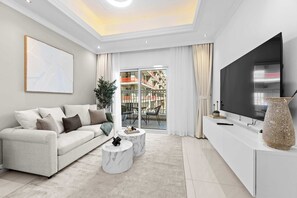 Ein modernes Wohnzimmer mit einer Couch, zwei Marmortischen, einem großen Fernseher und Blick auf einen Balkon mit Gartenmöbeln.