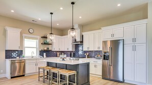 A modern kitchen with white cabinets, a stainless steel refrigerator, and a central island with stools. - Walk to Water & Greenway! Luxe Carolina Beach Home (Carolina Beach)