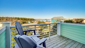 A wooden deck with a chair, overlooking a beach and a building with a blue roof. - Walk to Water & Greenway! Luxe Carolina Beach Home (Carolina Beach)