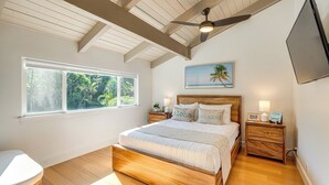 A bedroom with a wooden bed, a ceiling fan, a window with a view of greenery, and a wall-mounted TV. - Walk to Beach, Pool Access! 'Kahana Coastal Condo' (Lahaina)