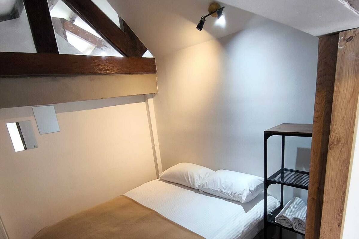 A small, single-bed room with a sloped ceiling, a wooden beam structure, a bed with a brown blanket, white pillows, and a metal shelf unit with folded towels.