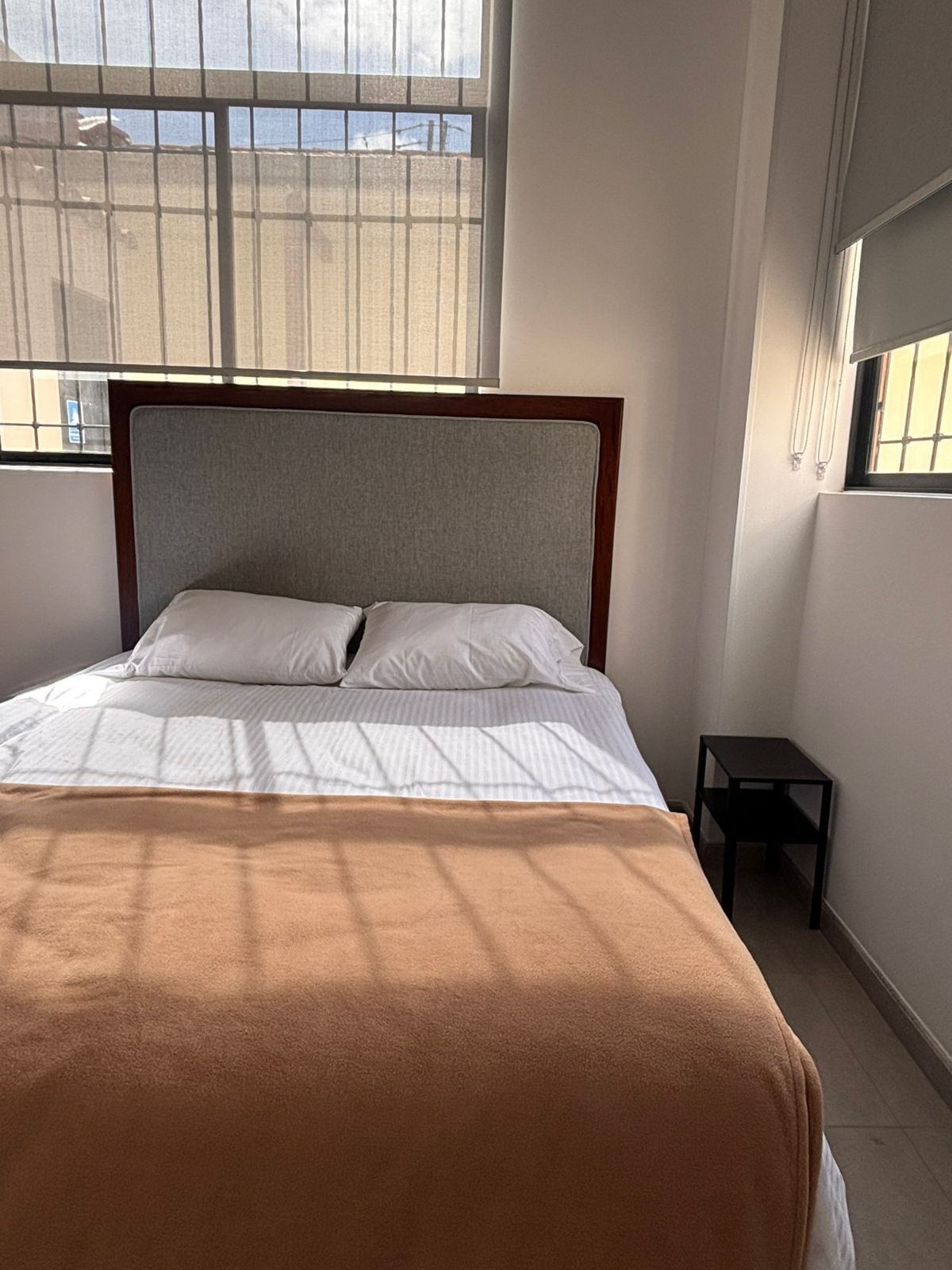 A bed with a headboard, a window with blinds, and a small table beside the bed.