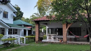 Una casa de dos pisos con techo verde, fachada blanca y antenas satelitales. Se observa un puente blanco que conduce a la casa, una estructura de ladrillo con techo rojo, y una hamaca.