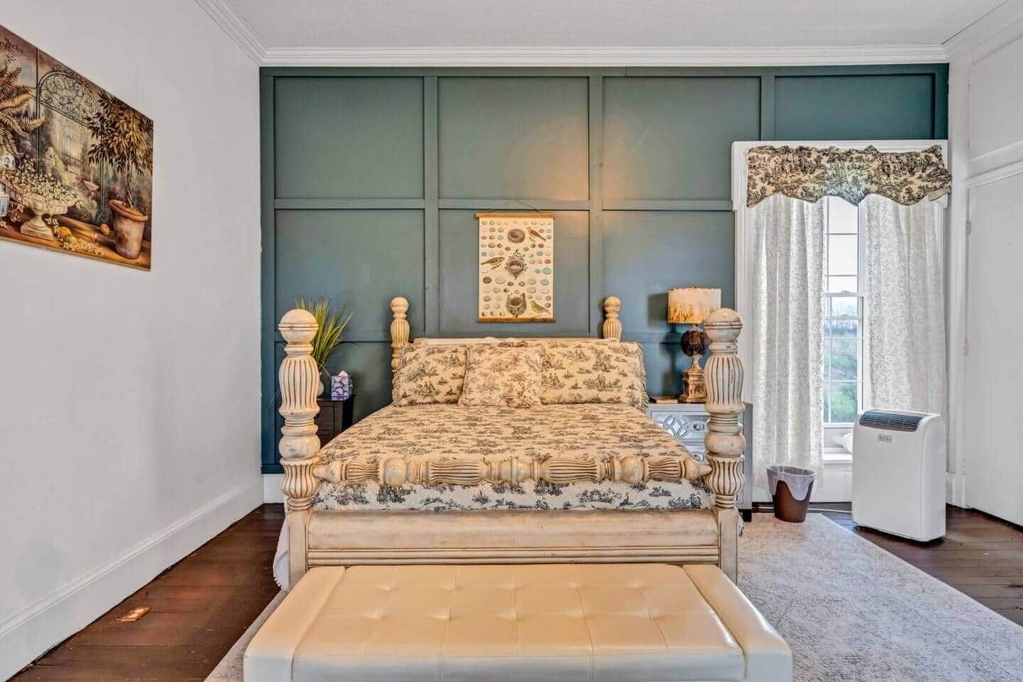A bedroom with a bed, a painting, a window with curtains, and a radiator.