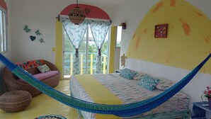 A room with a hammock, a bed with patterned bedding, a sofa with colorful cushions, and a view of greenery outside. - Colorful room very close to the beach. Only one lot behind the best beach | 2 (Holbox Island)