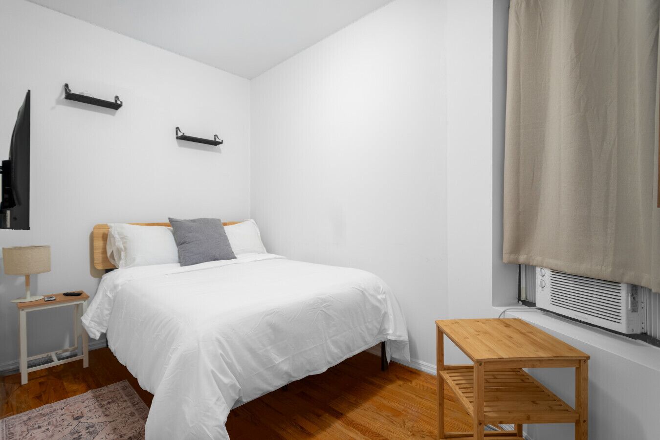 Un dormitorio con cama, mesitas de noche, una pequeña mesa de madera, un radiador y una ventana con cortinas.