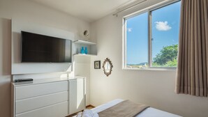 Ein Schlafzimmer mit Fernseher, Nachttisch, Bett, Fenster mit Vorhängen und Blick ins Freie.