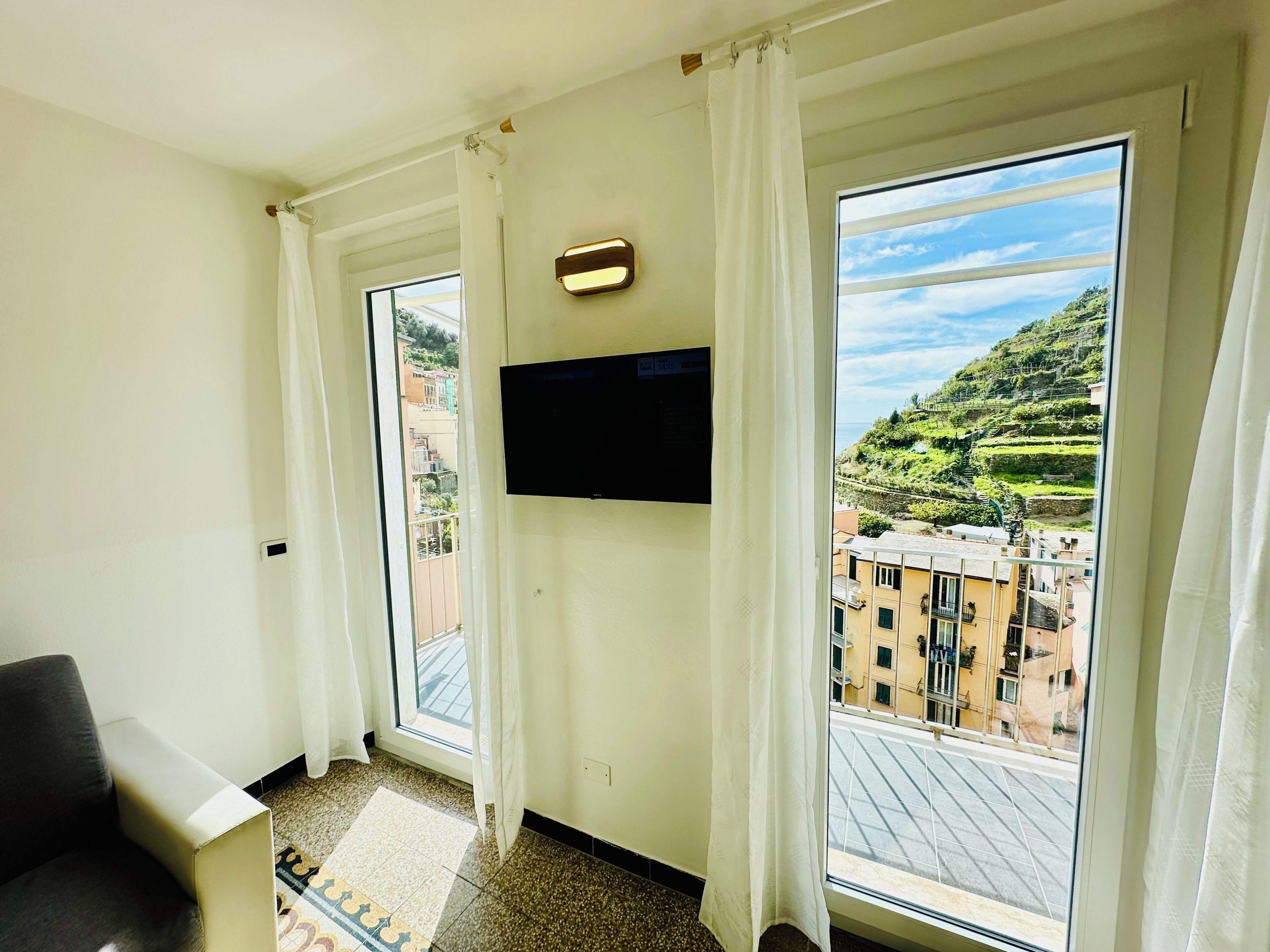 A room with a television mounted on the wall, a window with curtains, and a view of a hillside.