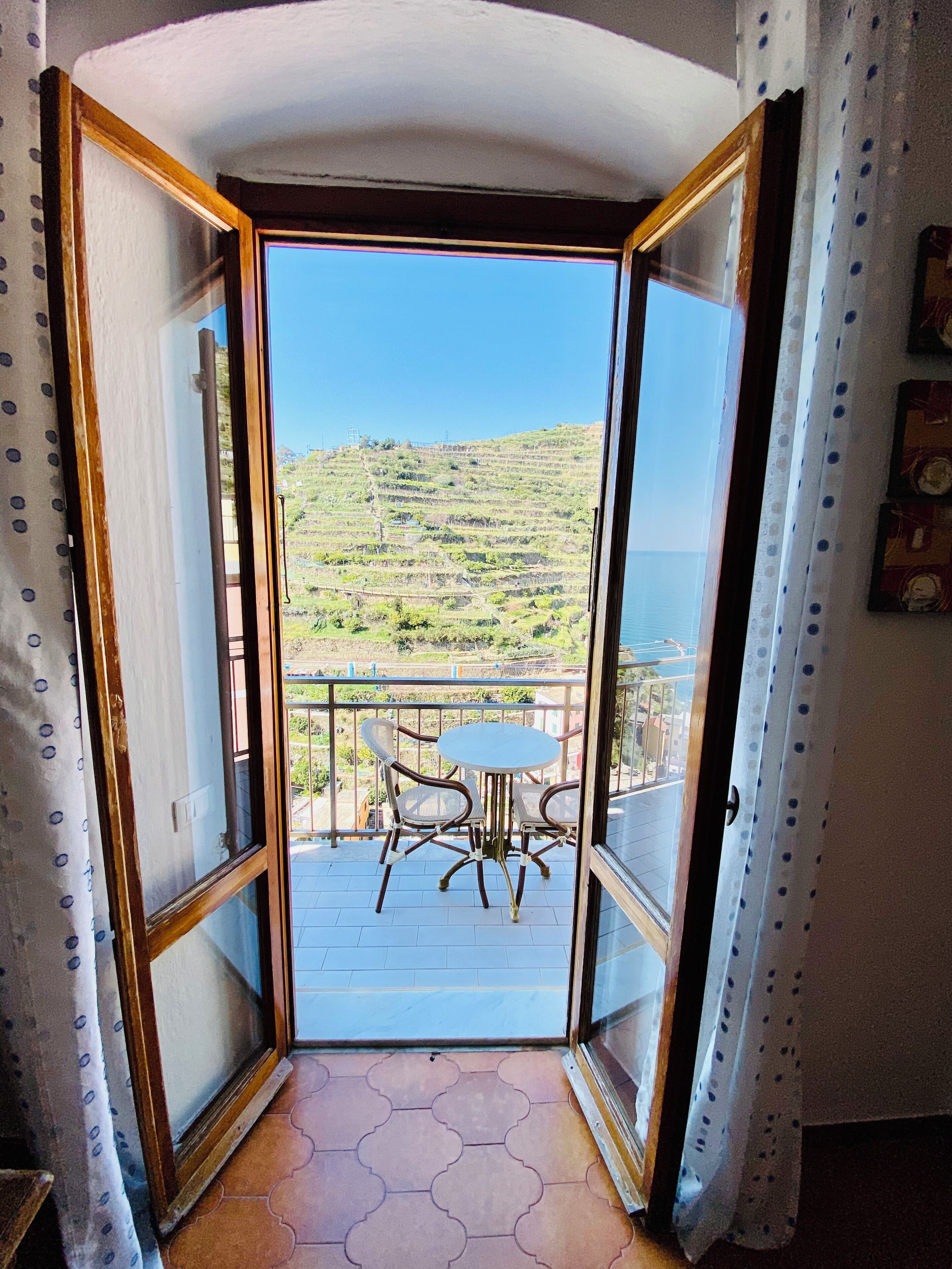 An open door leading to a balcony with a table and chairs, a view of a hillside, and a tiled floor.
