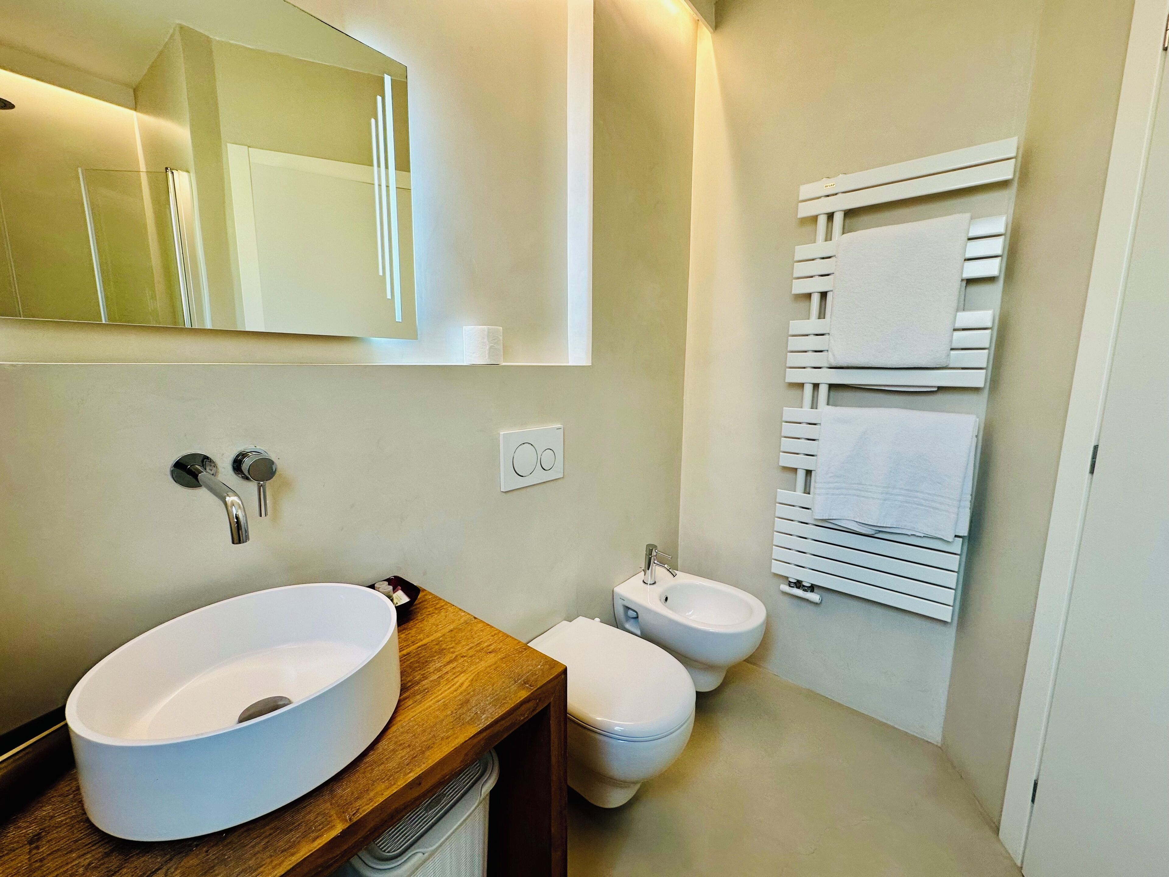 Ein modernes Badezimmer mit einem großen weißen Waschbecken, einer Toilette und einem Bidet. Es gibt einen Holzauszug, einen Spiegel und einen Handtuchhalter mit mehreren Handtüchern.