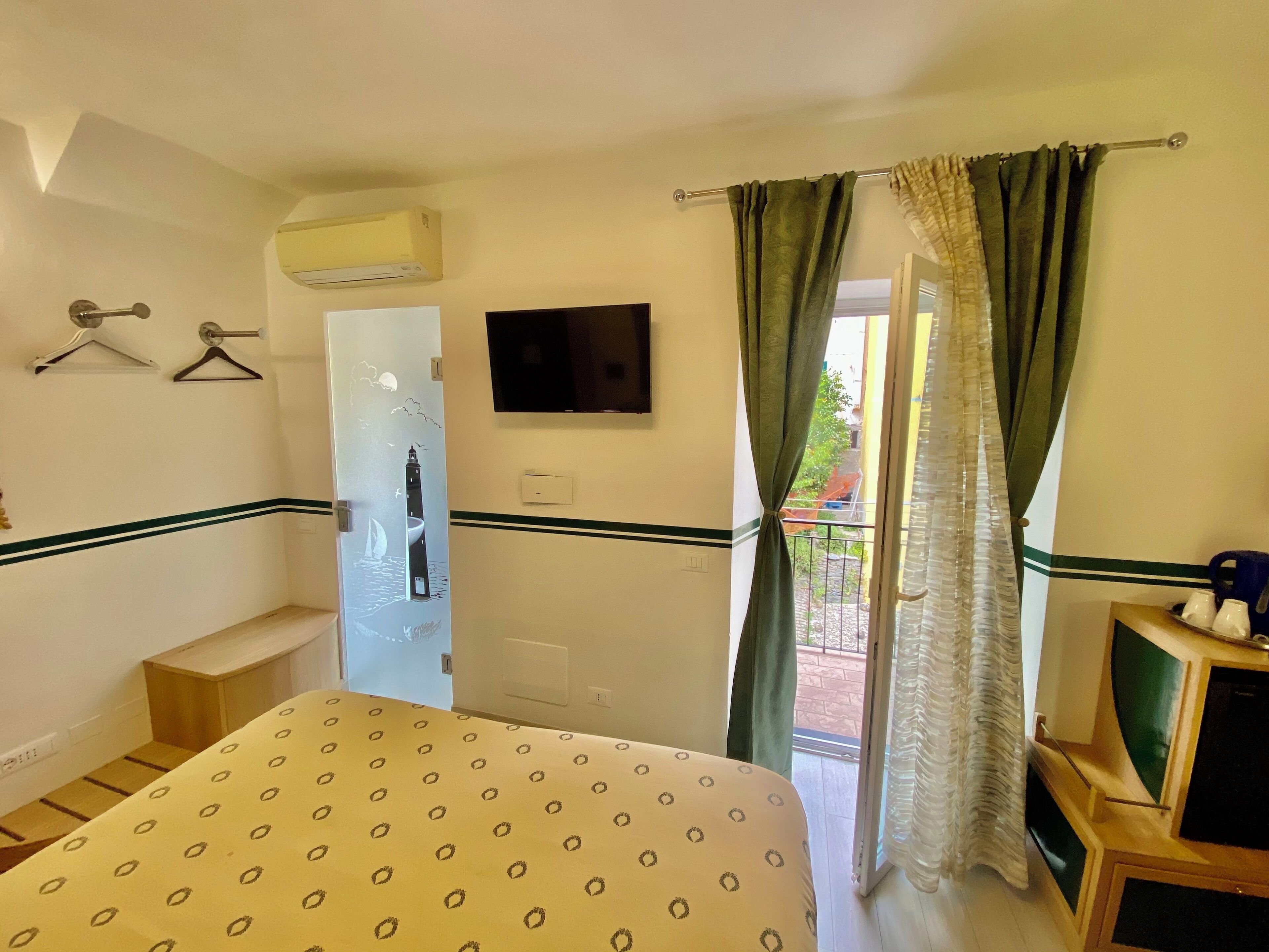 A hotel room with a bed, a TV, an air conditioning unit, a door with a view, and a balcony with curtains.