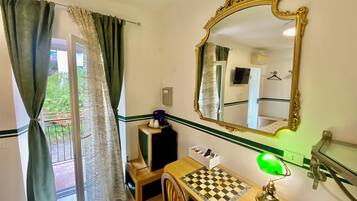 A room with a desk, a mirror, a chessboard, a lamp, and a view of the outside through a window with curtains.