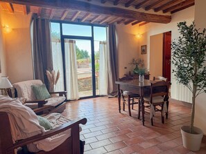Une pièce chaleureuse avec un lit, une table en bois et des chaises, une plante en pot, et une vue sur l’extérieur grâce à une porte vitrée.
