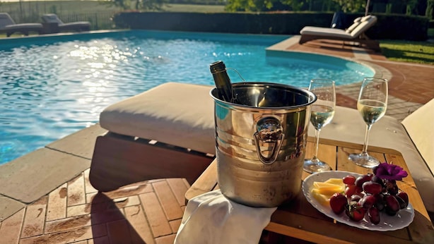 Une scène de bord de piscine avec un seau en métal, deux verres de vin blanc et une assiette de fruits.