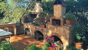 Une cuisine extérieure en pierre, dotée d’une cheminée, entourée de plantes en pot et agrémentée d’une table de patio.