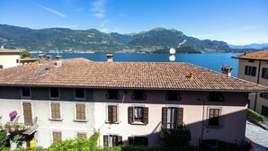 A building with a tiled roof, multiple windows with shutters, and a view of a lake and mountains in the background. - La Corte del Governo 565 (Lezzeno)