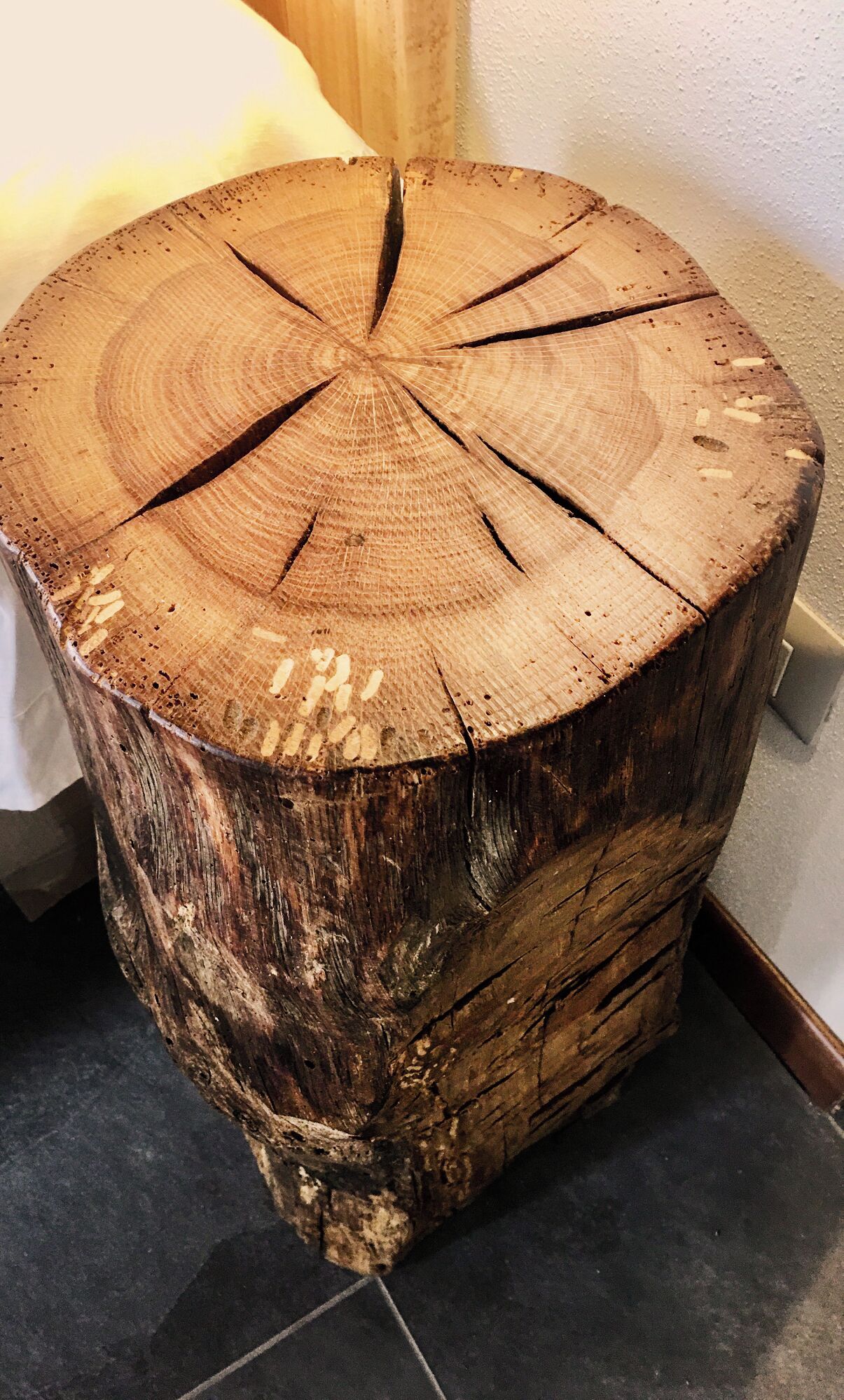 Een houten kruk met een zichtbaar patroon van jaarringen, geplaatst op een betegelde vloer in de buurt van een bed en een muur.