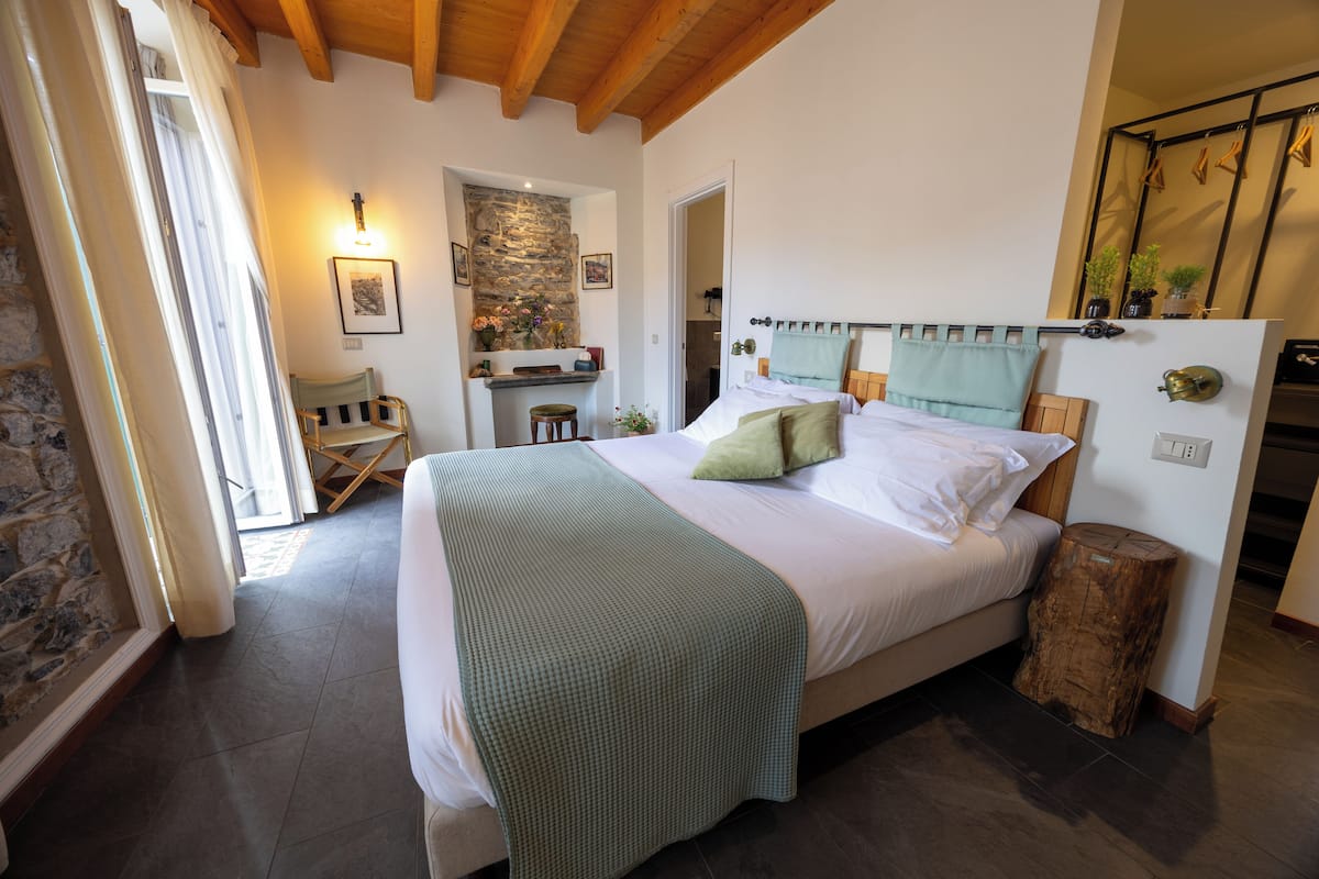 A bedroom with a large bed, a stone wall, a wooden ceiling, and a small stool.