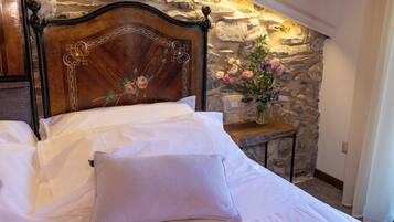 A bedroom with a stone wall, a wooden headboard, a white bed, and a small table with a vase of flowers.