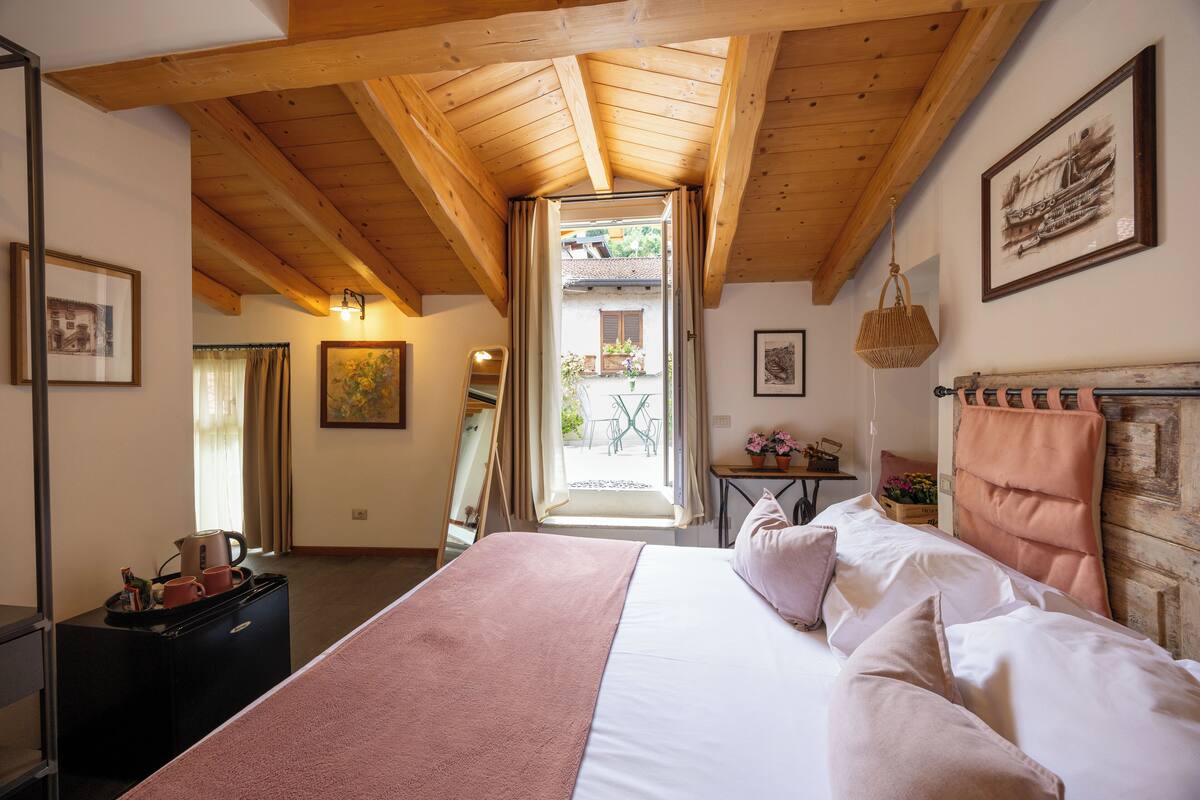 A bedroom with a wooden ceiling, a bed with white linens, a bedside table with a lamp, and a window with curtains.