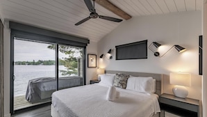 A bedroom with a bed, a nightstand, a wall-mounted lamp, a ceiling fan, and a large window overlooking a body of water.
