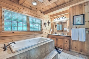 A wooden cabin bathroom with a large bathtub, wooden cabinets, a double sink, and a window with blinds. - 5 BR Luxury Log Cabin | Game Room + 2 Decks (Sapphire)