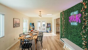 Un espace repas avec une table et des chaises en bois, un néon mural et, au fond, des jeux d’arcade.