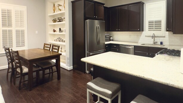 A modern kitchen with dark cabinets, a stainless steel refrigerator, and a dining area with a wooden table and chairs. - Best Location near Downtown/The Heights (Houston)