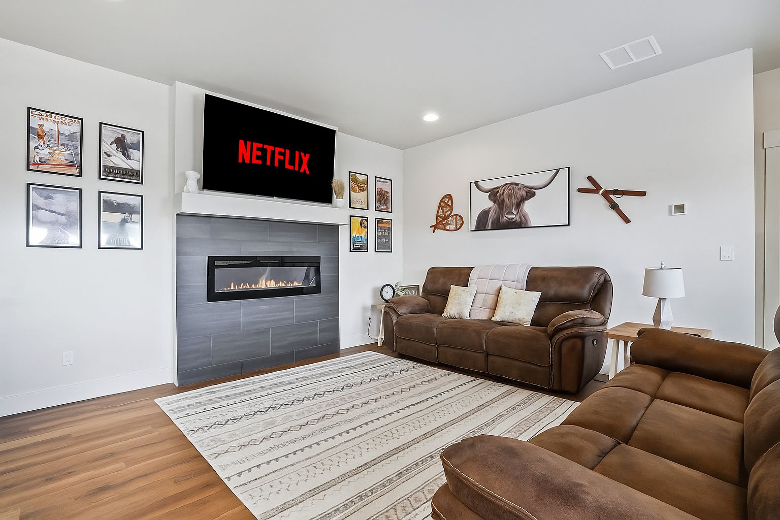 A modern living room with a fireplace, a brown leather sofa, a flat-screen TV displaying 'NETFLIX', and various framed pictures on the walls.