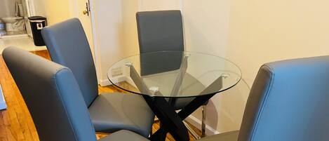 Una habitación con una mesa de vidrio rodeada por cuatro sillas azules, piso de madera y una puerta blanca con un interruptor de luz.