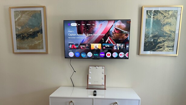 Ein an der Wand befestigter Fernseher mit einem Schreibtisch darunter, einem Spiegel und zwei gerahmten Kunstwerken.