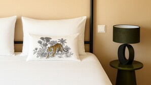 Un lit avec une literie blanche, un coussin à motifs de tigre et une lampe moderne posée sur une table de chevet.