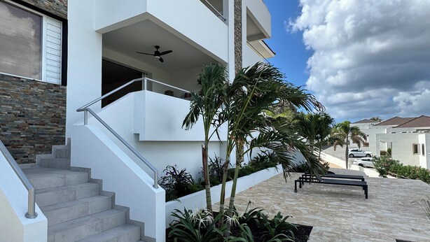 Une maison moderne à plusieurs étages, dotée d’un escalier menant à un balcon, et entourée de jardins aménagés et de palmiers.