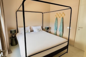 A four-poster bed with white bedding, two bedside tables with lamps, and a door to the right. - 2 bedroom apt @bluebay golf resort with pool. 5 min walk from bluebay beach (Willemstad)