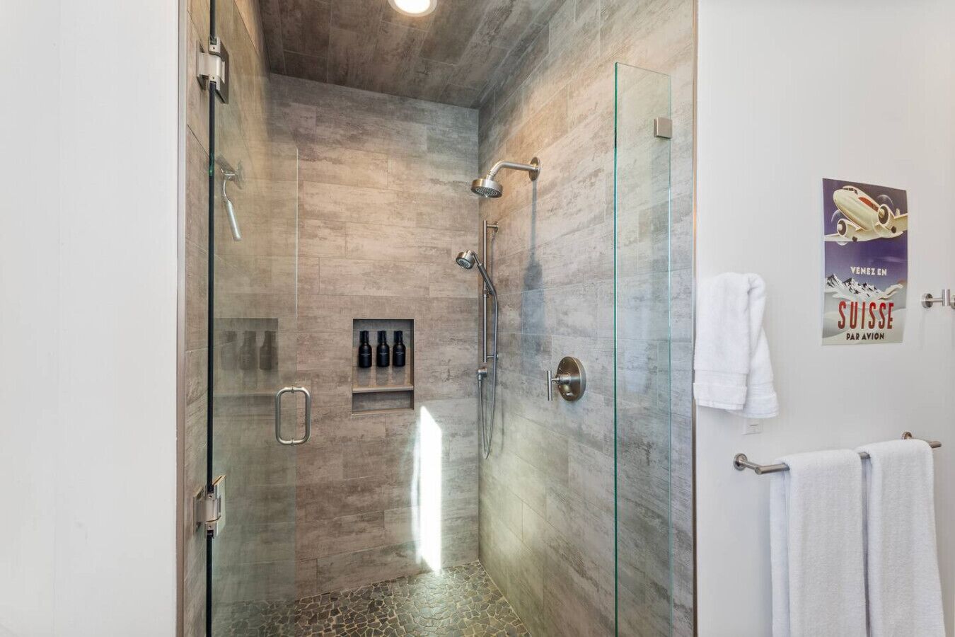 A modern bathroom with a glass shower enclosure, a built-in shelf with toiletries, and a poster on the wall.