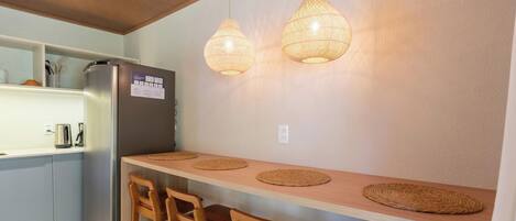 Una cocina con un refrigerador, sillas de madera, una mesa de madera y dos lámparas colgantes.