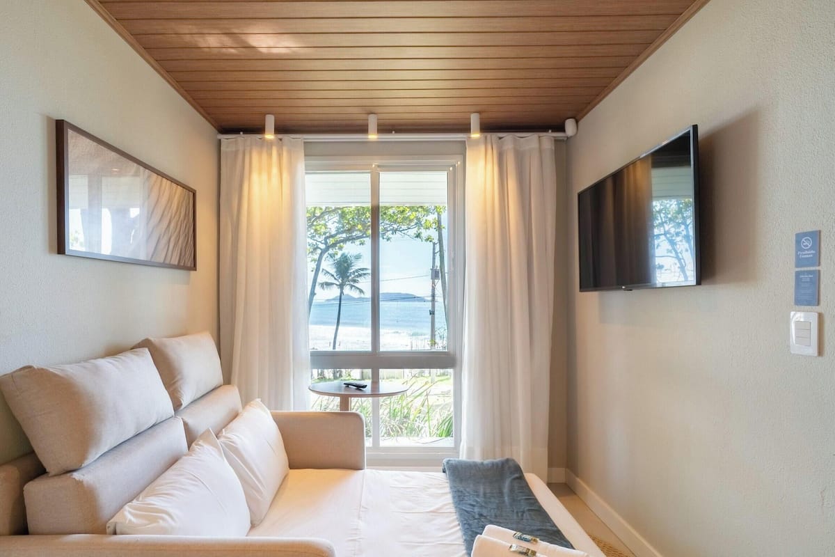 A hotel room with a wooden ceiling, a sofa with cushions, a TV mounted on the wall, and a window with curtains.