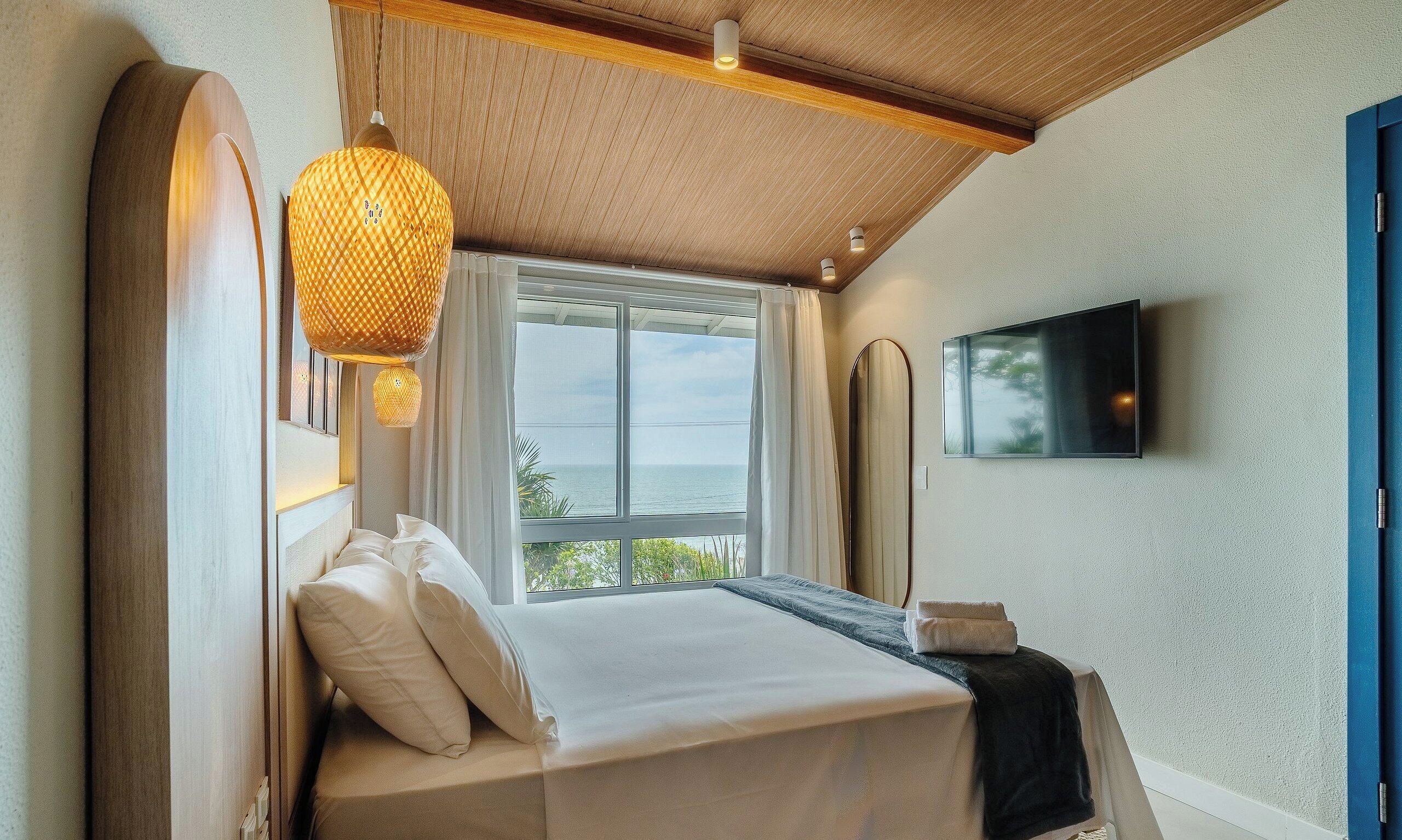 Una habitación de hotel con cama, un ventanal grande, televisor montado en la pared y techo con vigas de madera.