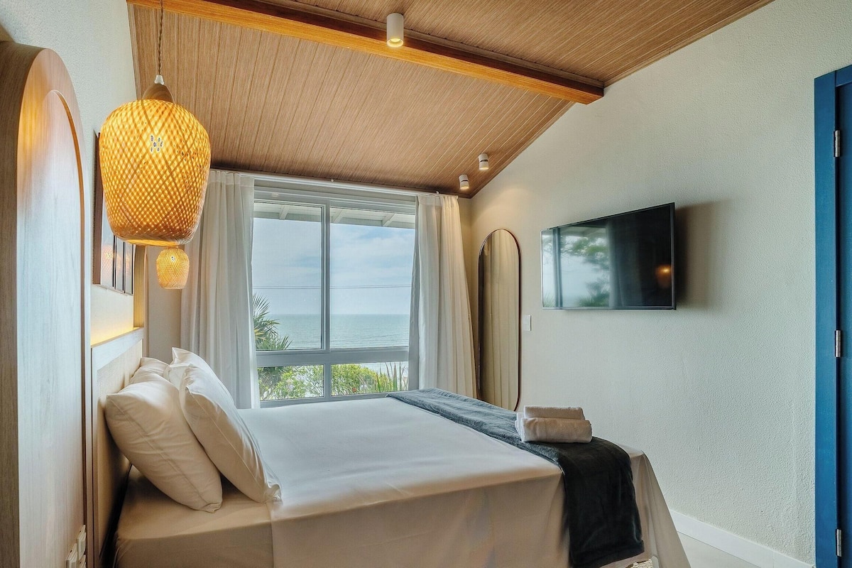 A bedroom with a bed, a television mounted on the wall, and a window with a view of the sea.