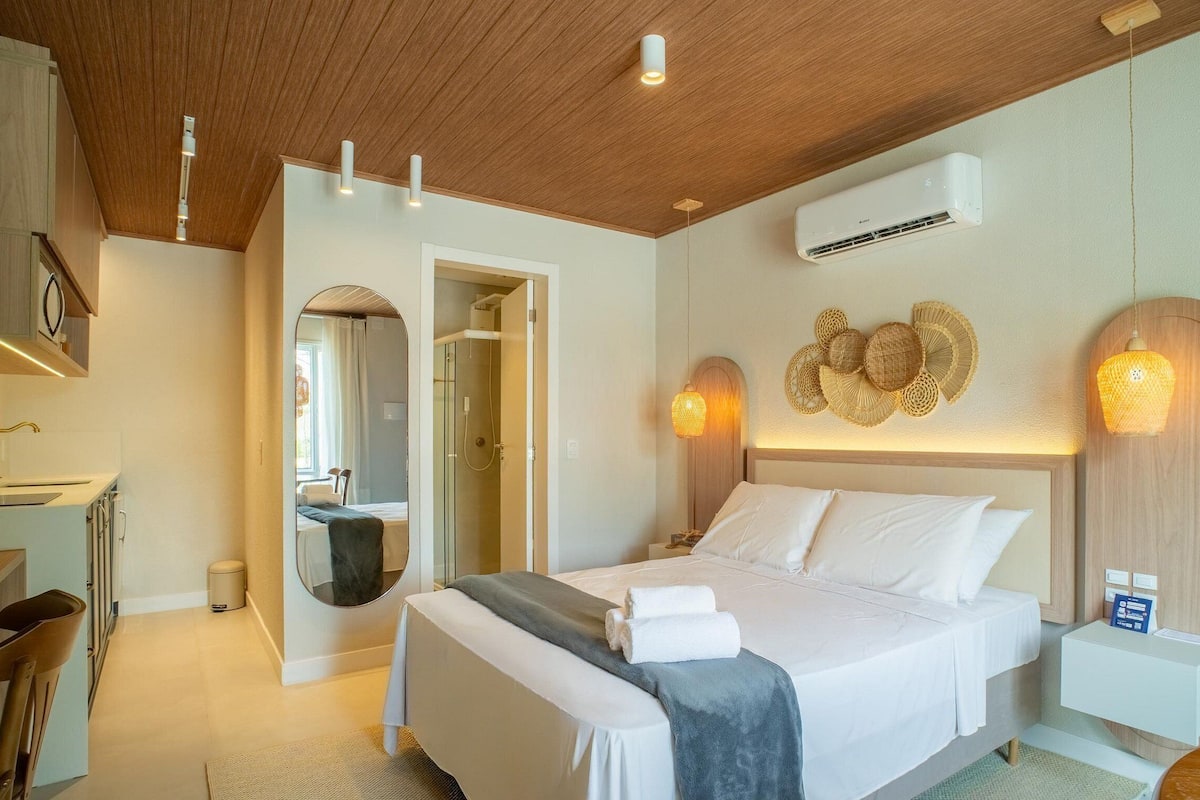A modern hotel room with a wooden ceiling, a bed with white linens, a round mirror, a shower, and a wall-mounted air conditioner.