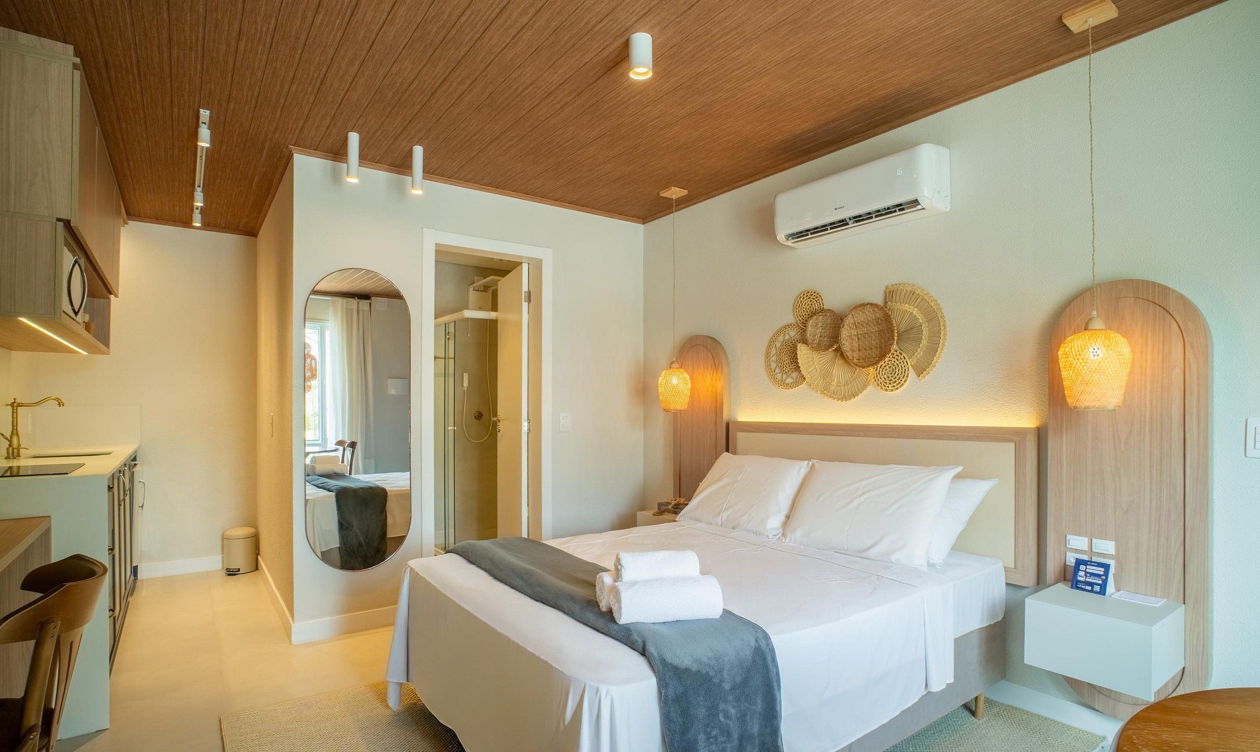 A modern hotel room with a wooden ceiling, a bed with white linens, a round mirror, a shower, and a wall-mounted air conditioner.