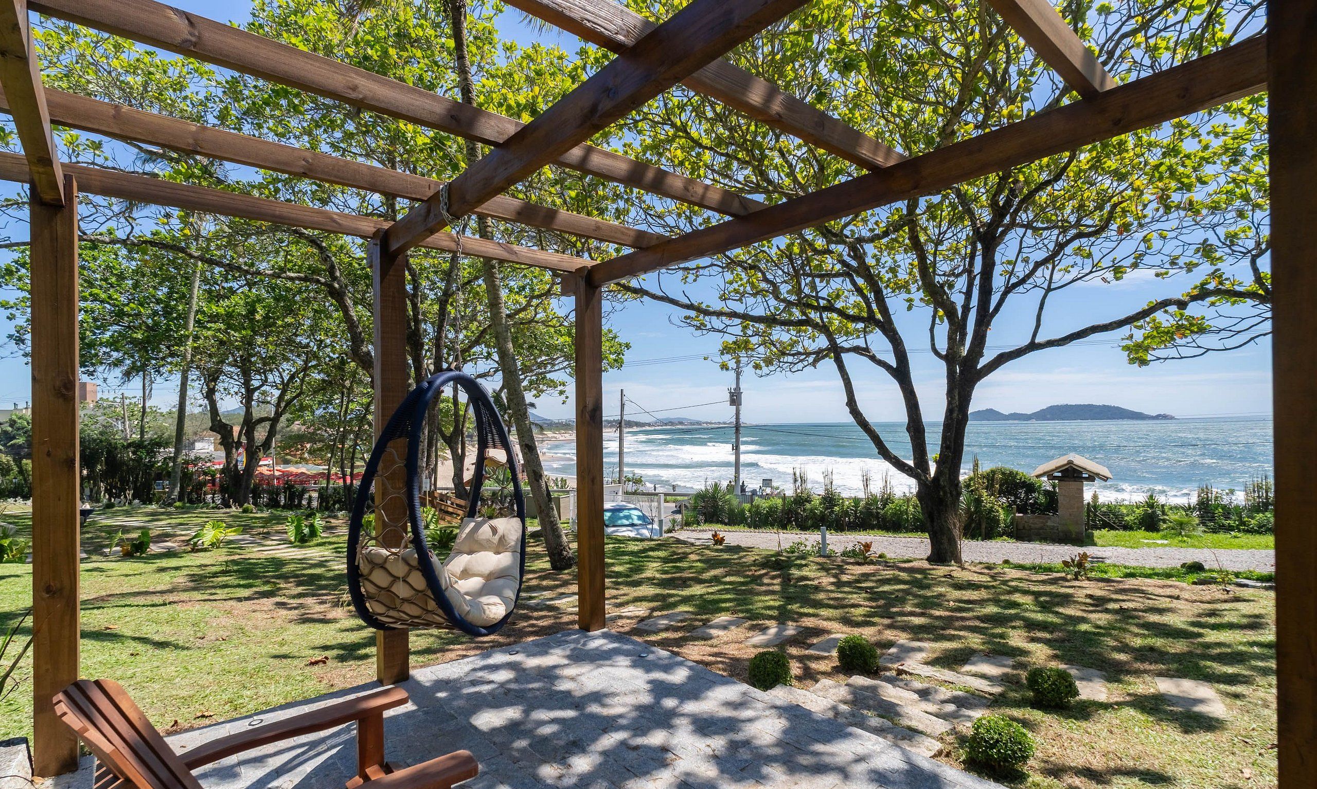 Una pérgola de madera con una silla colgante y un banco, con vista a una playa y al océano, con montañas al fondo.