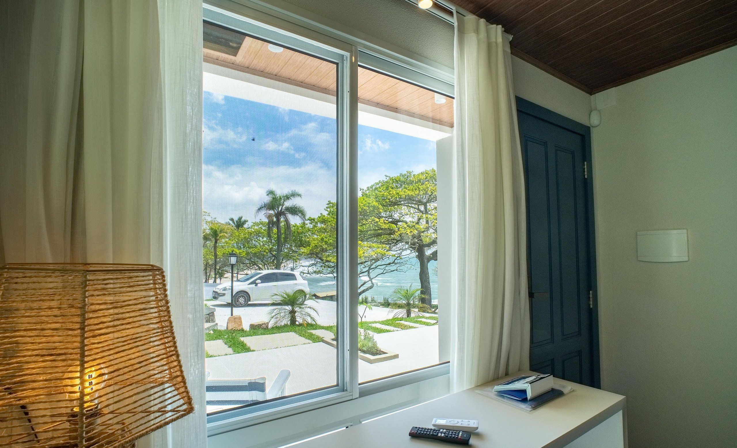 A room with a large window, a desk with a lamp, and a view of a parking area and greenery outside.