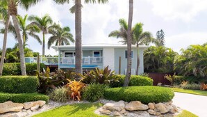 Una casa de dos pisos con balcón, rodeada de vegetación tropical y palmeras.