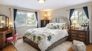 Une chambre à coucher avec un grand lit, deux tables de chevet avec des lampes, une commode, une chaise et une fenêtre avec des rideaux.