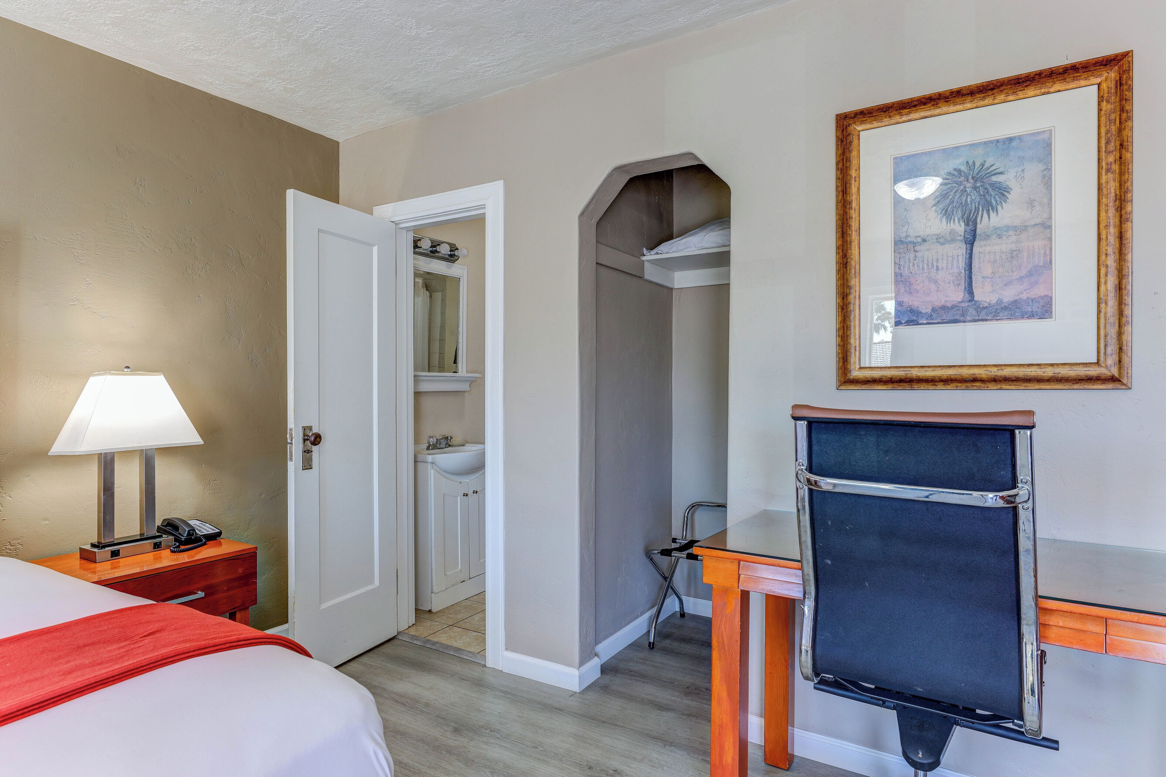 A hotel room with a bed, a desk, a chair, a lamp, a bathroom, and a framed picture on the wall.