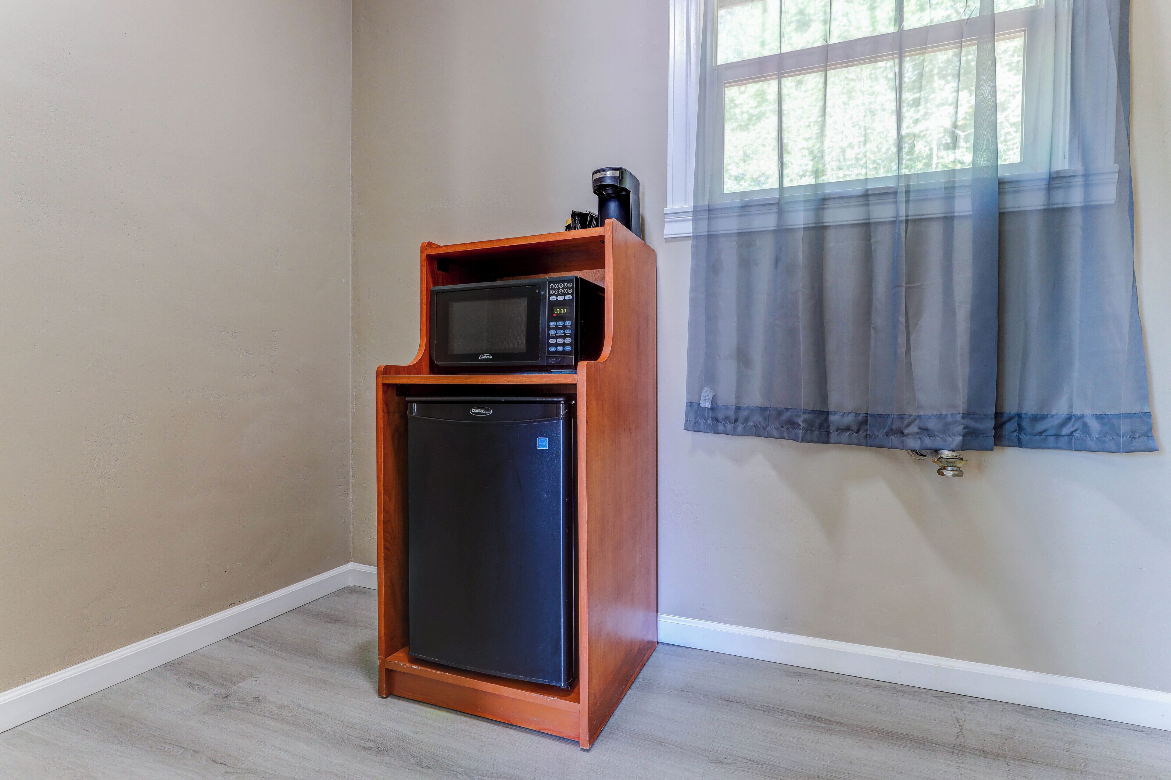 A compact kitchen area with a microwave and a small refrigerator, a window with a sheer curtain, and a light-colored wooden floor.
