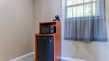 A compact kitchen area with a microwave and a small refrigerator, a window with a sheer curtain, and a light-colored wooden floor.